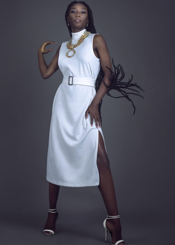 Locks of Beauty. Black female model with dreadlocks wearing white dress, high heels, and jewelry in grey studio portrait.