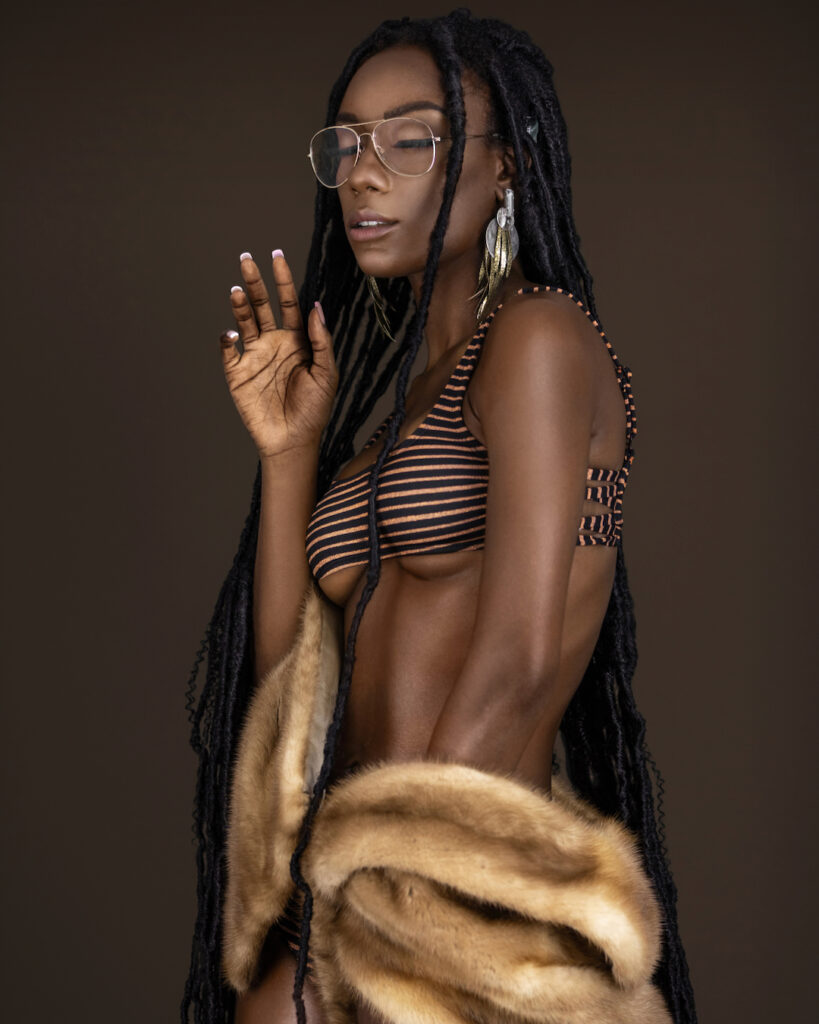Portrait of a serene young black female with long dreadlocks, beautiful makeup and moist lips posing by herself in a studio with dark background wearing jewelry, a bikini & fur coat.