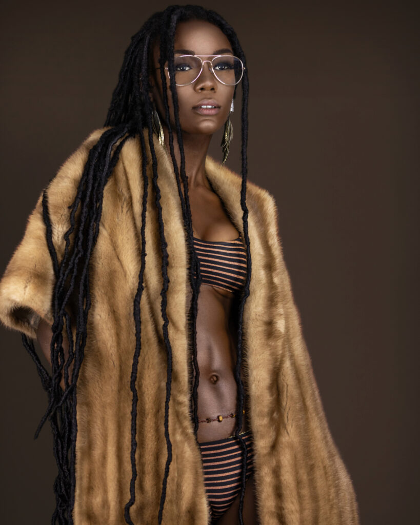 Portrait of a sexy young black female with long dreadlocks, beautiful makeup and moist lips posing by herself in a studio with dark background wearing jewelry, a bikini & fur coat.