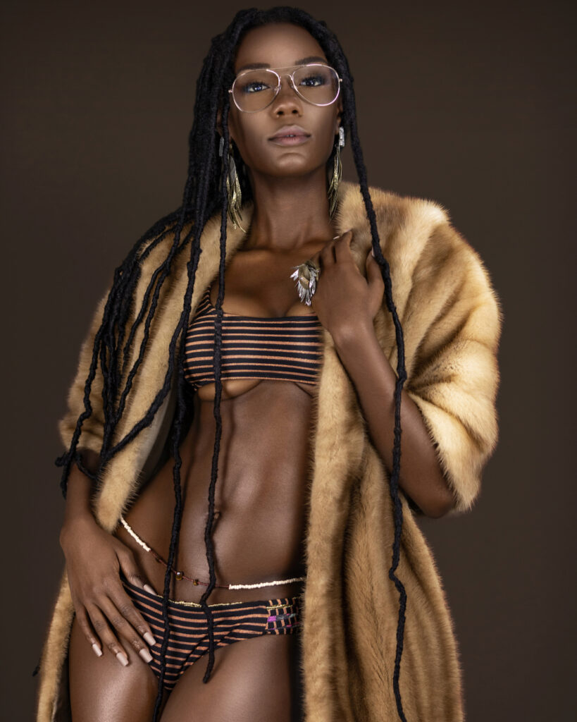 Portrait of an avantgardist young black female with long dreadlocks, beautiful makeup and moist lips posing by herself in a studio with dark background wearing jewelry, a bikini & fur coat.
