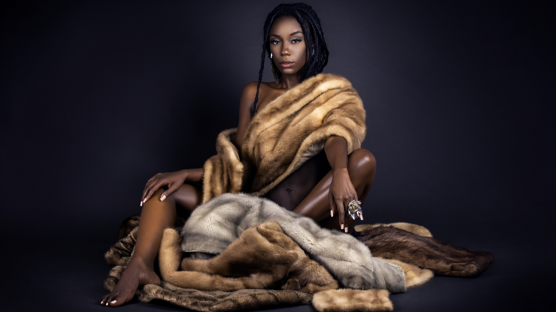 Portrait of a serene young black female with long dreadlocks, beautiful makeup and perfect lips sitting by herself on fur in a studio with dark background wearing jewelry & a fur coat.