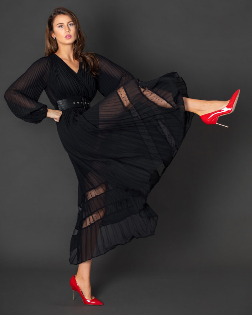 Portrait of a confident young white woman with wavy hair and beautiful makeup posing by herself inside a studio with a grey background wearing a black dress with a belt and red high heels.