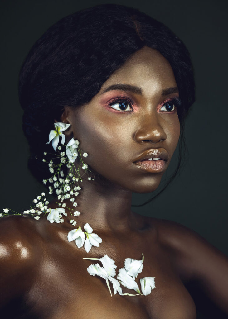The Ethereal Goddess. Portrait of a sensual young black female with curly long black hair, beautiful makeup and soft lips posing by herself in a studio with dark background wearing white flowers on her ears & chest.