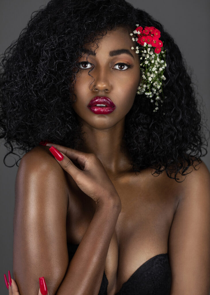 The Ethereal Goddess. Portrait of a sensual young black female with curly long black hair, beautiful makeup and red lips posing by herself in a studio with grey background wearing flowers in her hair.