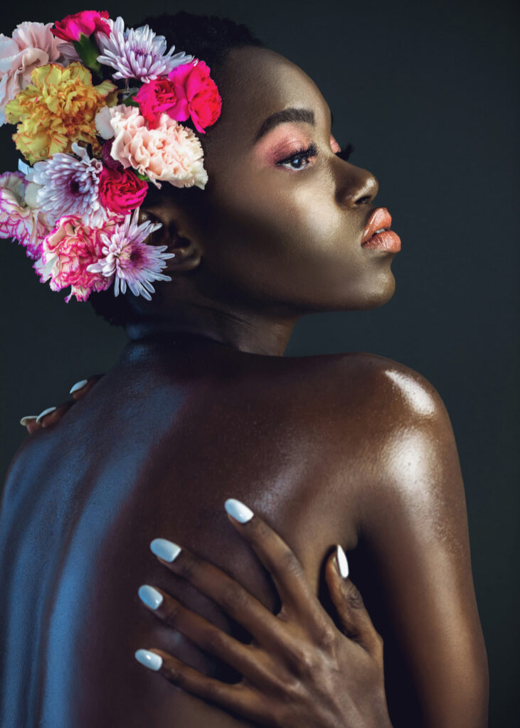 Portrait of a serene young black female with a short Afro, beautiful makeup and moist lips posing by herself in a studio with dark background wearing a bouquet of flowers in her hair.