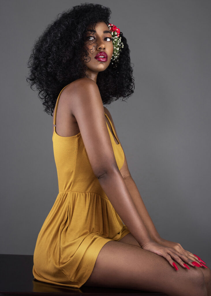 Portrait of a serene young black female with curly long black hair, beautiful makeup and red lips sitting on a desk by herself in a studio wearing a yellow dress & flowers in her hair.