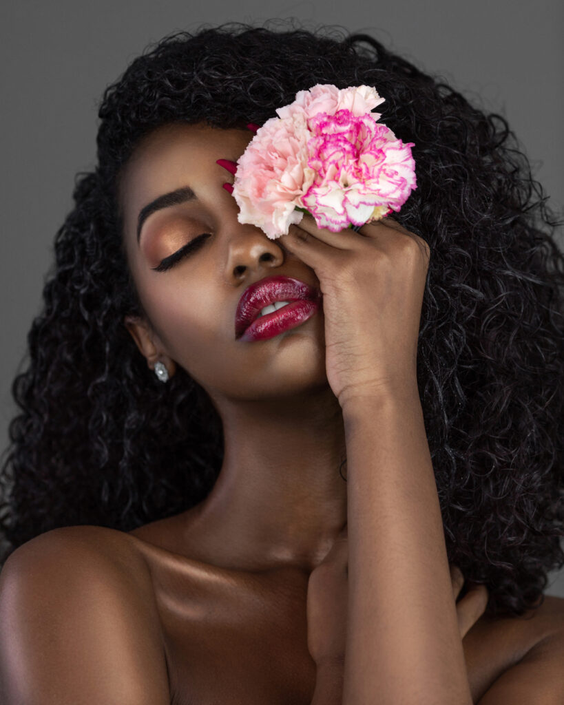 The Ethereal Goddess. Closeup portrait of a sensual young black female with curly long black hair, beautiful makeup and red lips posing by herself in a studio covering one eye with a beautiful flower.