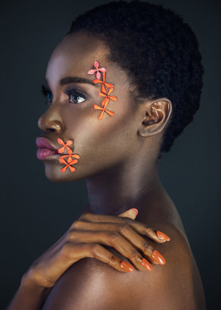 Portrait of a serene young black female with a beautiful short Afro, beautiful makeup, purple lips & orange nails posing by herself in a studio with dark background wearing orange flowers on her face.