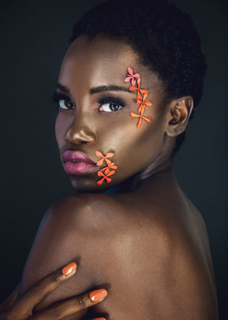 The Ethereal Goddess. Portrait of a sexy young black female with a beautiful short Afro, beautiful makeup, purple lips & orange nails posing by herself in a studio with dark background wearing orange flowers on her face.