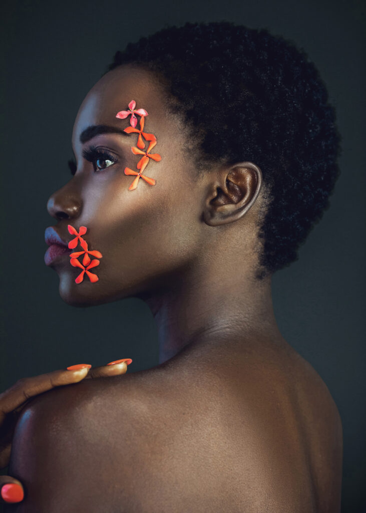 Portrait of a sensual young black female with a beautiful short Afro, beautiful makeup, purple lips & orange nails posing by herself in a studio with dark background wearing orange flowers on her face.