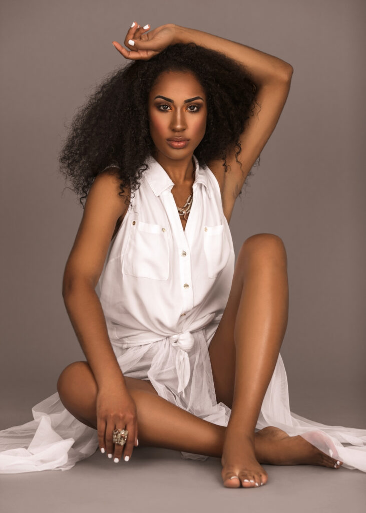 Young Latin woman with curly hair in a white dress sitting barefoot in studio for Curls Poppin editorial.