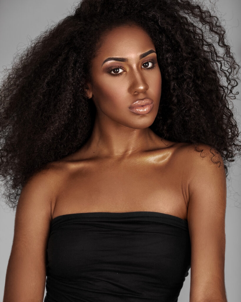 Closeup portrait of a confident young Latin woman with long curly hair and beautiful makeup posing by herself inside a studio with a grey background wearing a black wrapped top and white shorts.