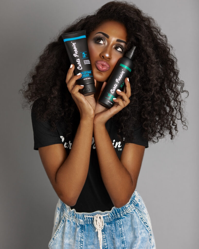 Closeup portrait of a lovely young Latin woman with long bouncy curly hair and beautiful makeup posing by herself inside a studio with a grey background holding a bottle of hair product in each hand.
