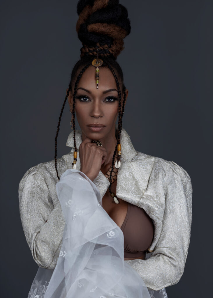 Portrait of a sensual Black woman with Updo black hair and beautiful makeup sitting by herself inside a studio with grey background wearing white jacket with brown bikini top and jewelry.