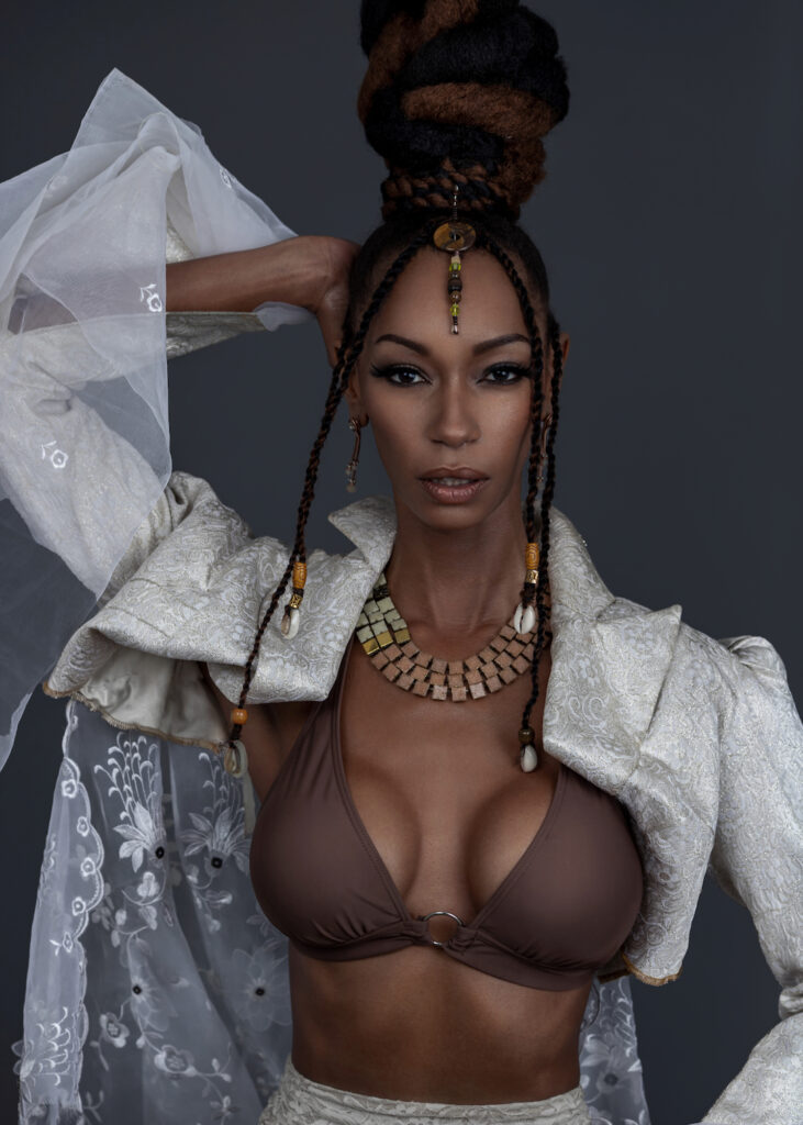 Portrait of a confident Black woman with Updo black hair and beautiful makeup sitting by herself inside a studio with grey background wearing white jacket with brown bikini top and jewelry.