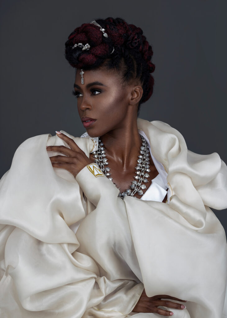 The Exhibitionist fashion editorial. Waist up portrait of a serene Black woman with Updo black hair and beautiful makeup sitting by herself inside a studio with grey background wearing white jacket and jewelry.