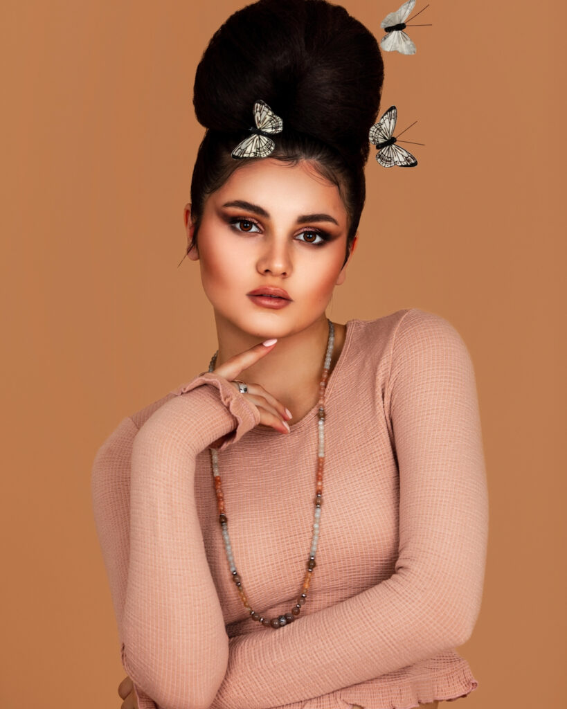Portrait of a confident Latin girl with long wavy hair in an up-do and beautiful makeup posing by herself surrounded by butterflies inside a studio with pecan color background wearing a pink top.