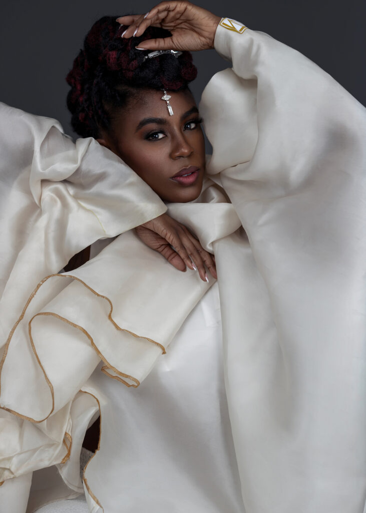 Waist up portrait of a sensual Black woman with Updo black hair and beautiful makeup sitting by herself inside a studio with grey background wearing white jacket and jewelry.
