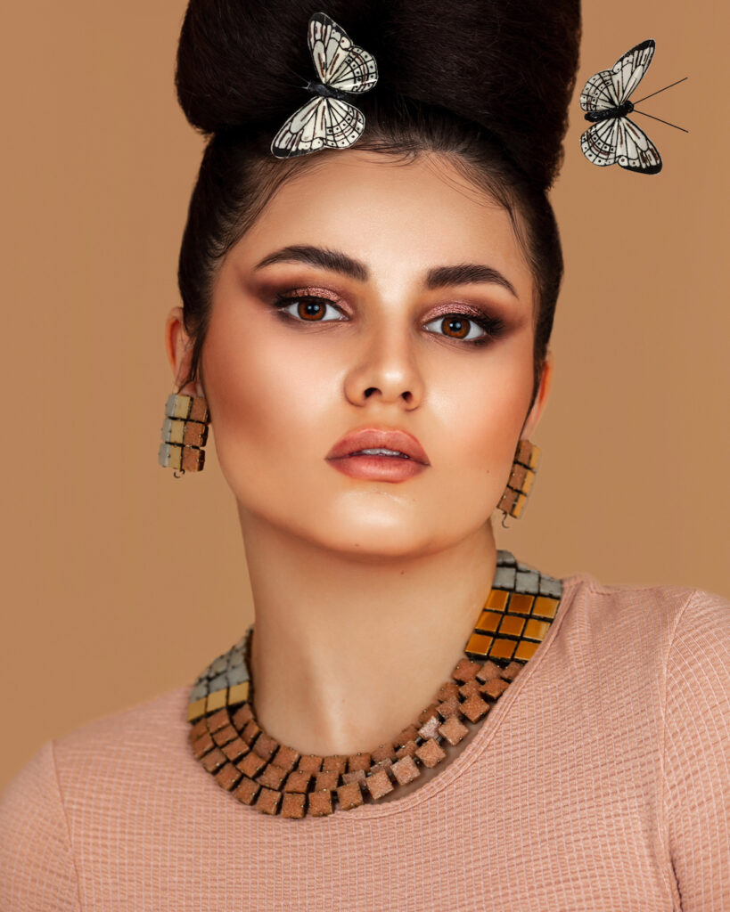 Close-up portrait of a sensual Latin girl with long wavy hair in an up-do and beautiful makeup posing by herself surrounded by butterflies inside a studio with pecan color background wearing jewelry.