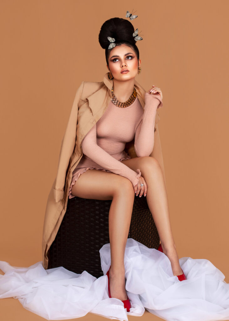 Portrait of a sexy Latin girl with long wavy hair in an up-do and beautiful makeup sitting by herself surrounded by butterflies inside a studio with pecan color background wearing a short pink outfit.