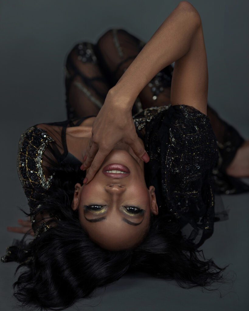 Portrait of a sensual Black woman with short black hair and beautiful makeup laying by herself inside a studio with grey background wearing black pants with blouse.