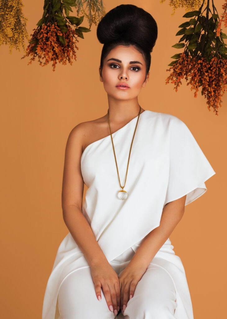 Portrait of a lovely Latin girl with long wavy hair in an up-do, beautiful makeup posing by herself surrounded by flowers inside a studio with pecan color background wearing a white evening dress.