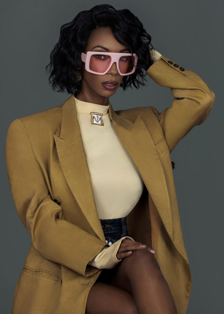 Portrait of a sexy Black woman with short black hair and beautiful makeup sitting by herself inside a studio with grey background wearing mustard colored suit jacked with big pink shades.