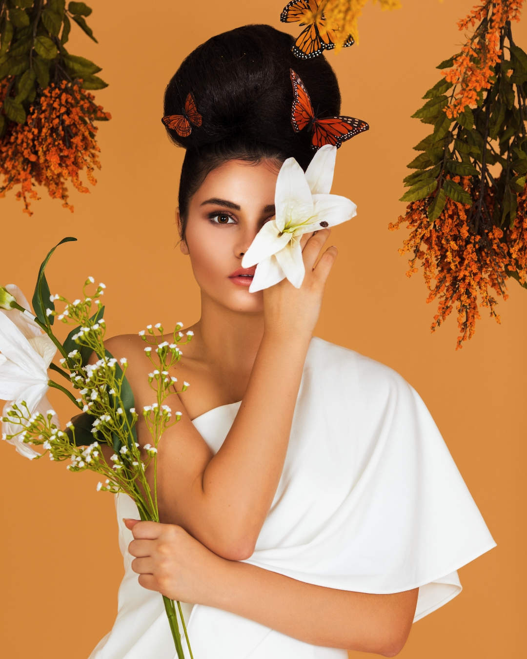 Fashion magazine editorial. Portrait of a shy Latin girl with long wavy hair in an up-do, beautiful makeup posing by herself surrounded by flowers inside a studio with pecan color background wearing a white evening dress.