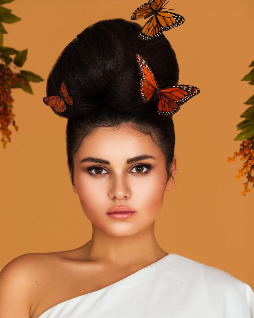 Portrait of a lovely Latin girl with long wavy hair in an up-do and beautiful makeup posing by herself surrounded by flowers and butterflies inside a studio with pecan color background.