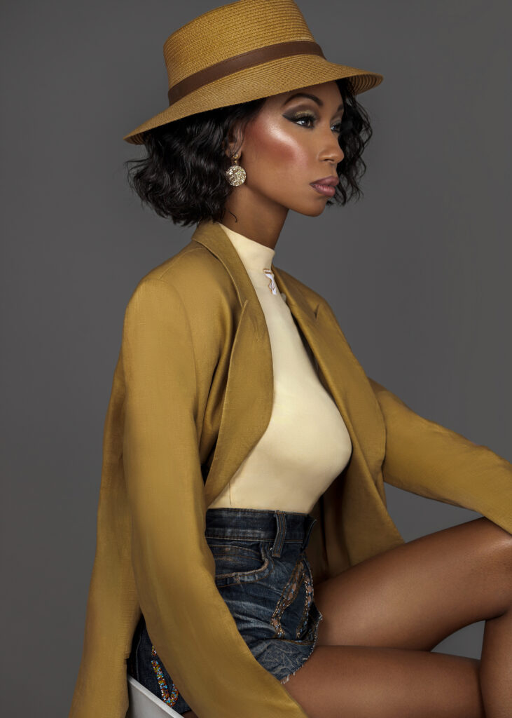 The Exhibitionist fashion editorial. Portrait of a confident Black woman with short black hair and beautiful makeup sitting by herself inside a studio with grey background wearing mustard colored suit jacked with a fedora hat.
