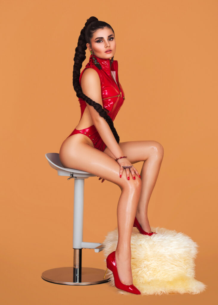 Portrait of a sexy Latin girl with very long ponytail hair, beautiful makeup sitting by herself on a bar chair inside a studio with pecan color background wearing a red leather body and high heels.