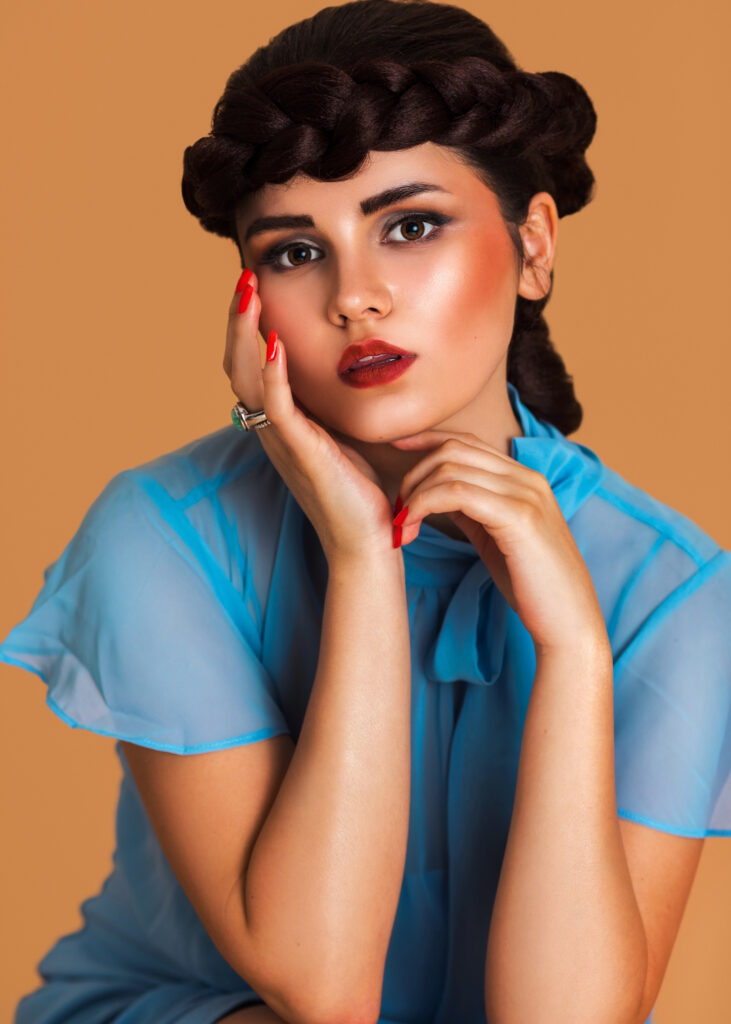 Closeup portrait of a lovely Latin girl with long braided hair and beautiful makeup sitting by herself on a bar chair inside a studio with pecan color background wearing a blue summer dress.