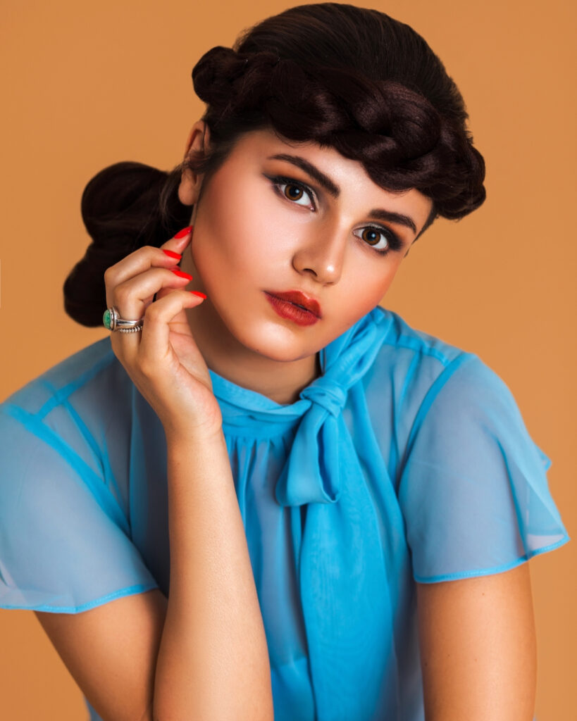 Closeup portrait of a beautiful Latin girl with long braided hair and beautiful makeup sitting by herself on a bar chair inside a studio with pecan color background wearing a blue summer dress.