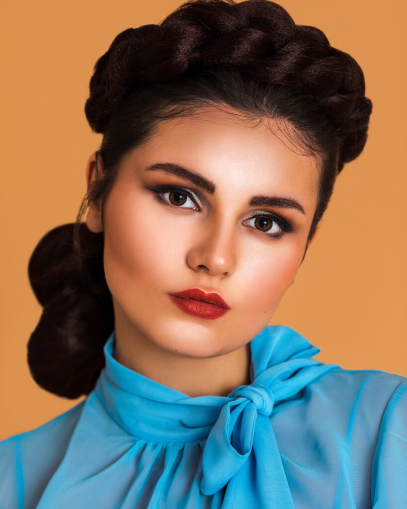 Closeup portrait of a gorgeous Latin girl with long braided hair and beautiful makeup sitting by herself on a bar chair inside a studio with pecan color background wearing a blue summer dress.