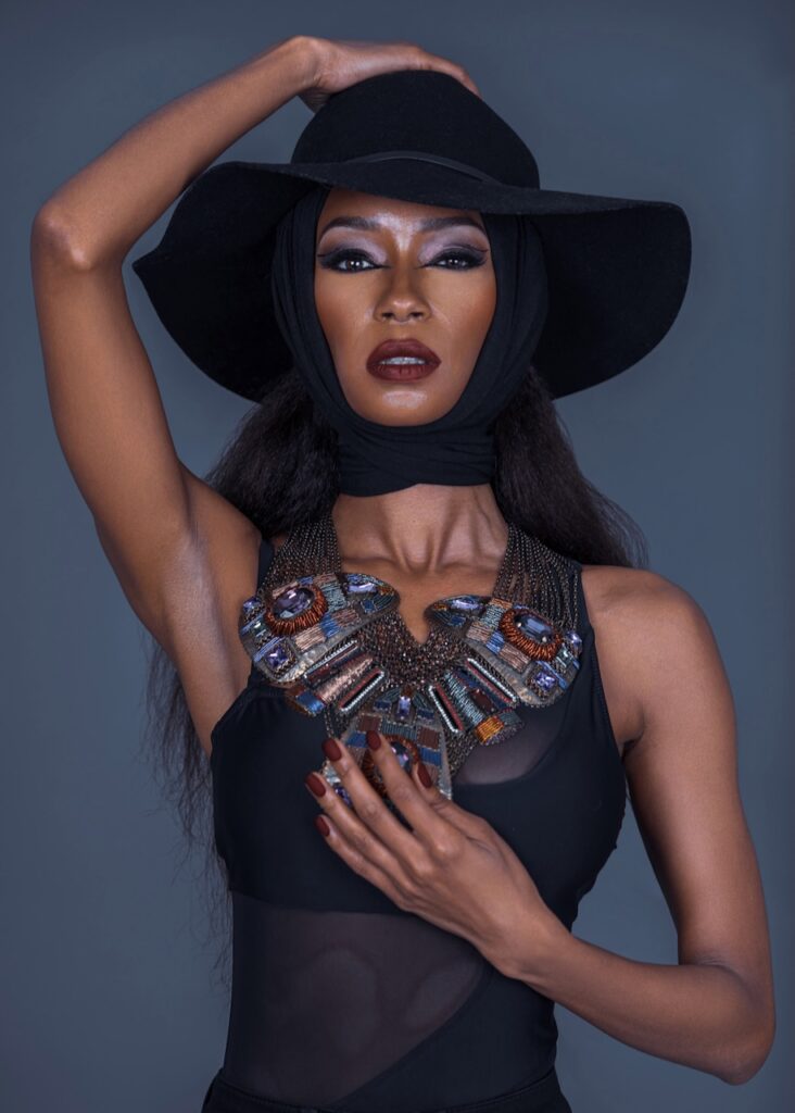 Fashion editorials 2025. Portrait of confident Black woman with long curly black hair and beautiful makeup standing by herself inside studio with grey background wearing jewelry necklace and a black hat.