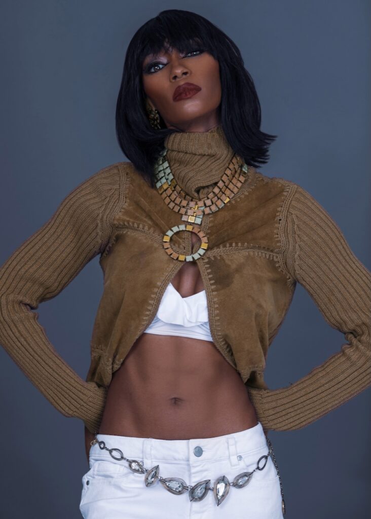 Fashion editorials 2025. Portrait of confident Black woman with straight black hair and beautiful makeup standing by herself inside studio with grey background wearing jewelry necklace and brown sweater. 