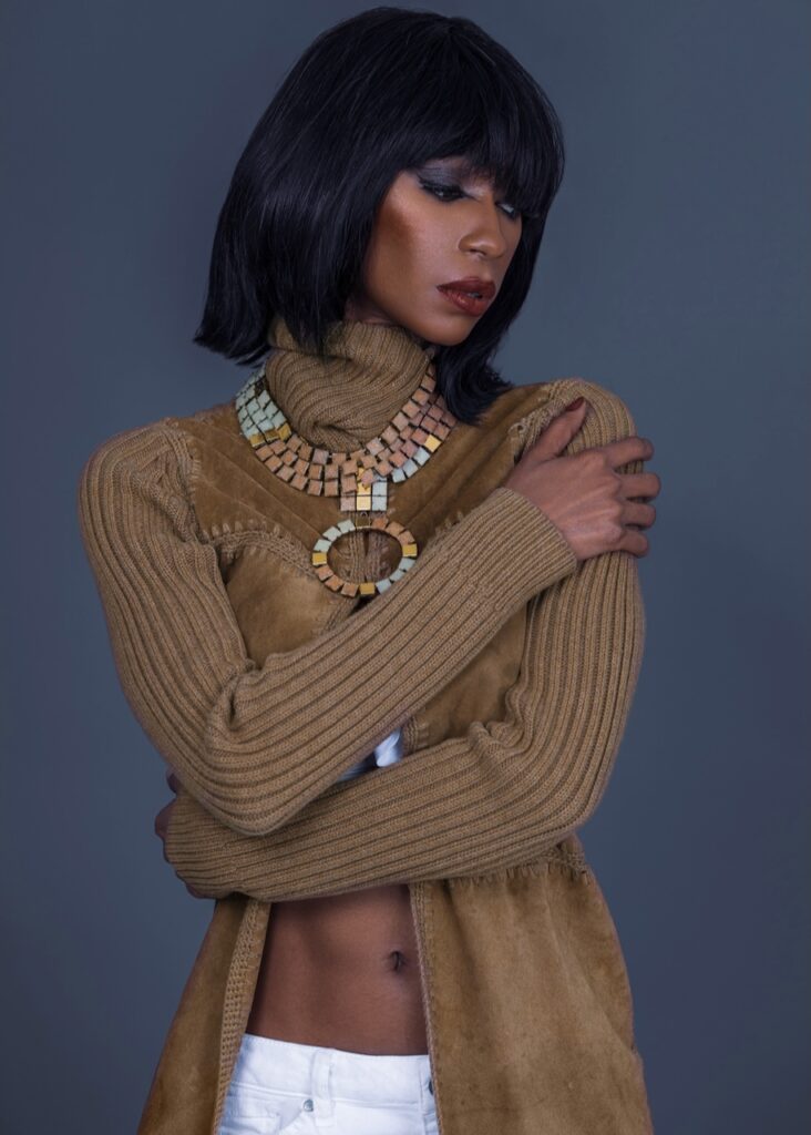 Portrait of serene Black woman with straight black hair and beautiful makeup standing by herself inside studio with grey background wearing jewelry necklace and brown sweater.