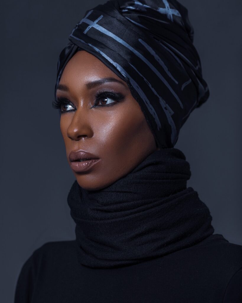 Fashion editorials 2025. Closeup portrait of serene Black woman with beautiful makeup standing by herself inside studio with grey background wearing blue silk turban and black sweater.