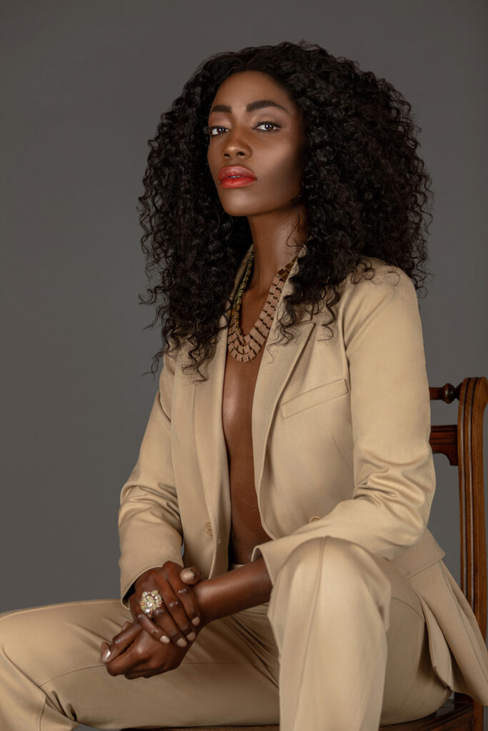 Portrait of a sensual young black woman with long curly black hair and beautiful makeup sitting by herself on a wooden chair in a studio with a grey background wearing a beige suit and jewelry.
