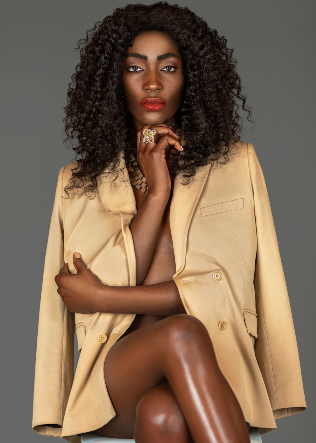Spring Is In The Air. 3/4 portrait of a happy black woman with long curly black hair and beautiful makeup sitting by herself in a studio with gray background just wearing a beige jacket and jewelry.