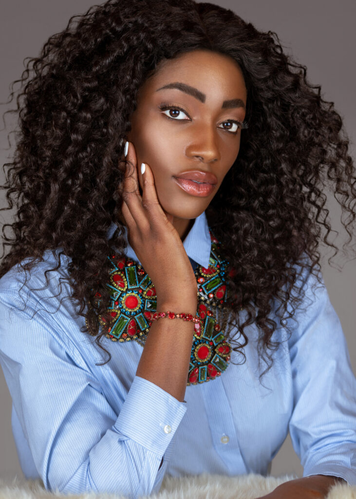 Portrait of a beautiful young black woman with long curly black hair, beautiful makeup sitting by herself on fur in a studio with a grey background wearing a blue shirt and colorful jewelry.