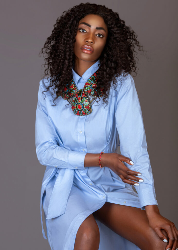 Portrait of a serene young black woman with long curly black hair and beautiful makeup sitting on a bar chair by herself in a studio with a grey background wearing a blue dress and colorful jewelry.