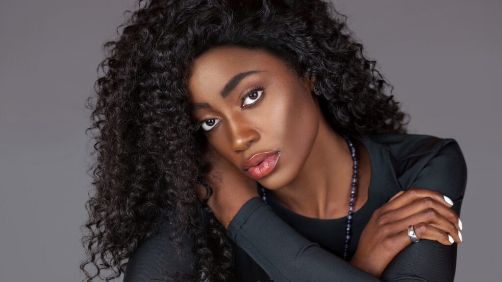 Spring Is In The Air. Black woman in long-sleeve black shirt and jewelry sitting on fur with grey background.