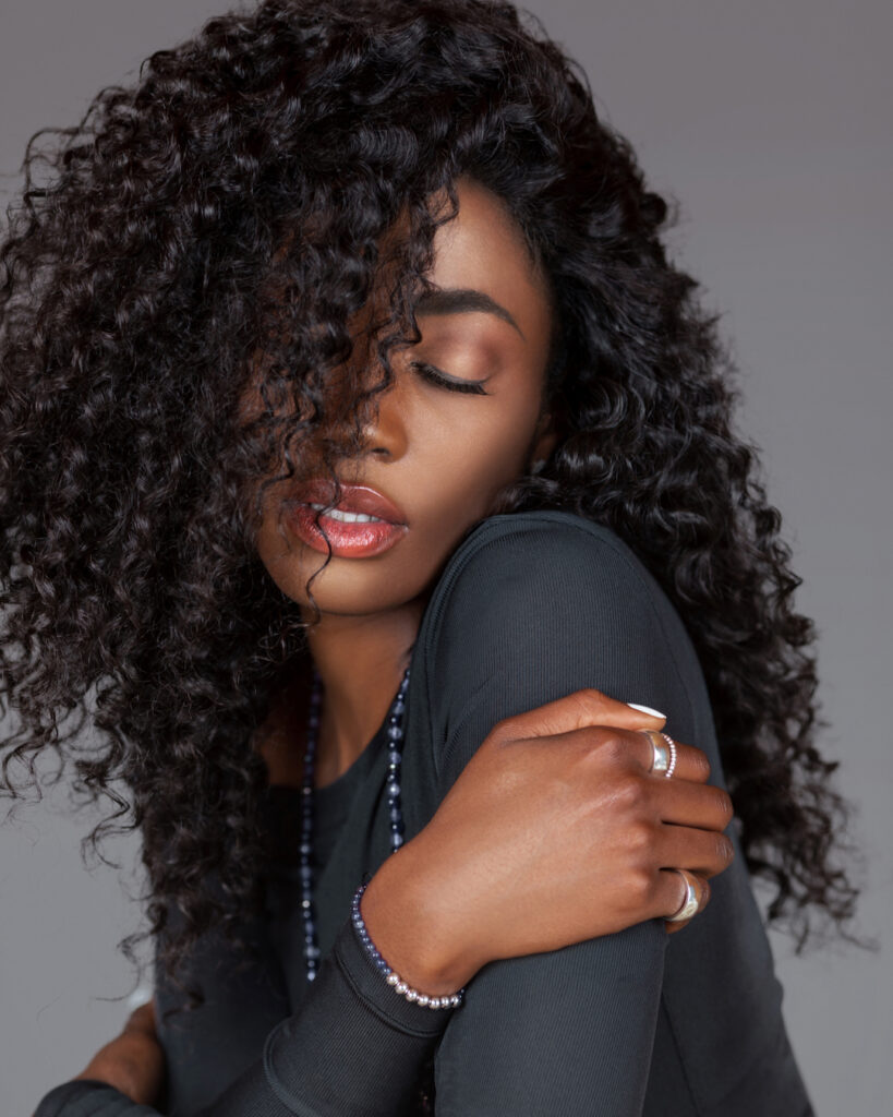 Portrait of a sensual young black woman with long curly black hair, beautiful makeup sitting by herself on fur in a studio with a grey background wearing a black long sleeve shirt and jewelry.