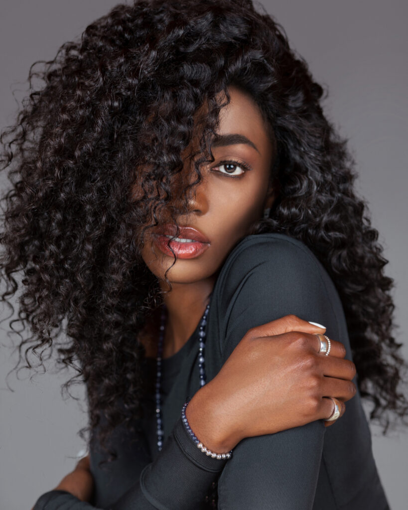 Portrait of an attractive young black woman with long curly black hair, beautiful makeup sitting by herself on fur in a studio with a grey background wearing a black long sleeve shirt and jewelry.