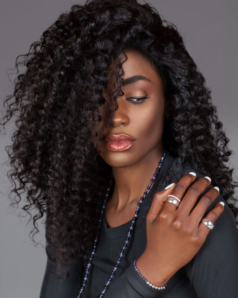 Portrait of a serene young black woman with long curly black hair, beautiful makeup sitting by herself on fur in a studio with a grey background wearing a black long sleeve shirt and jewelry.