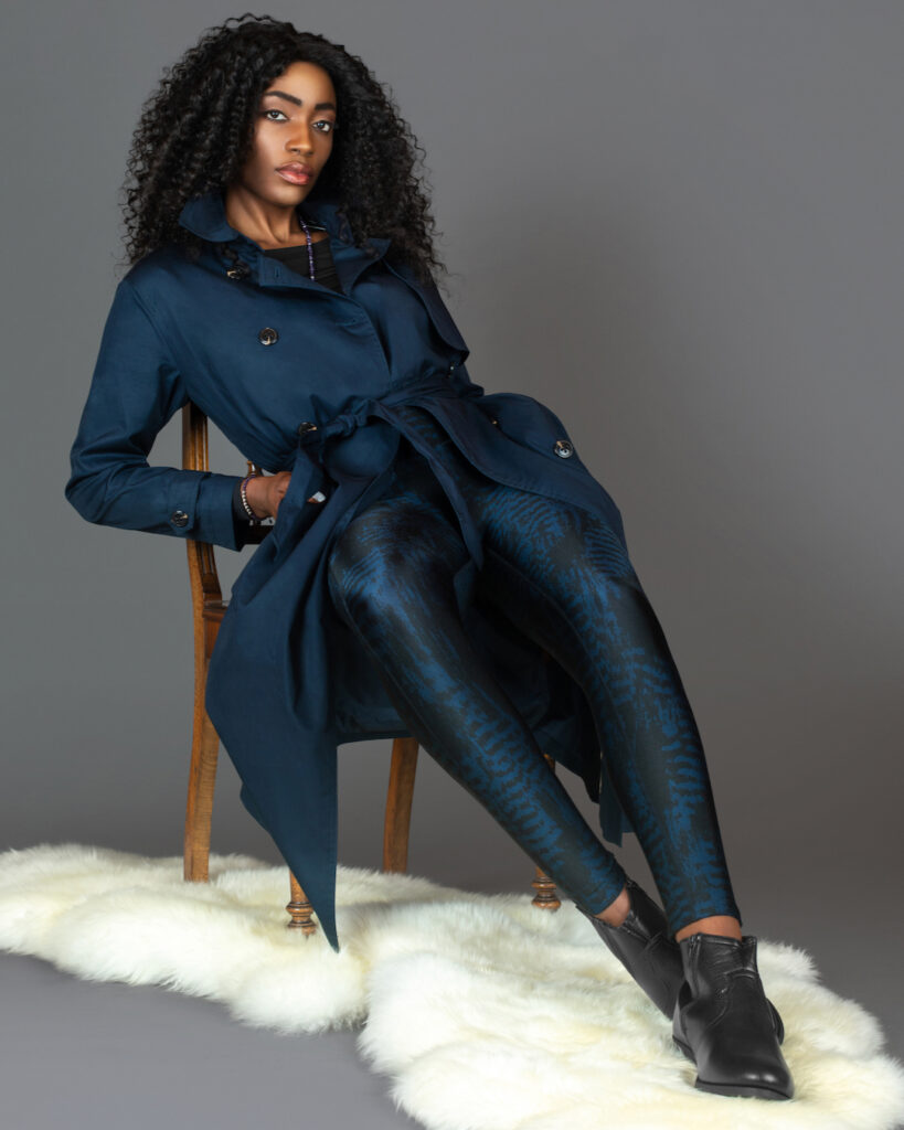 Portrait of a sexy black woman with long curly black hair, beautiful makeup sitting by herself on fur in a studio with a grey background wearing a blue trench coat and leggings with black boots.