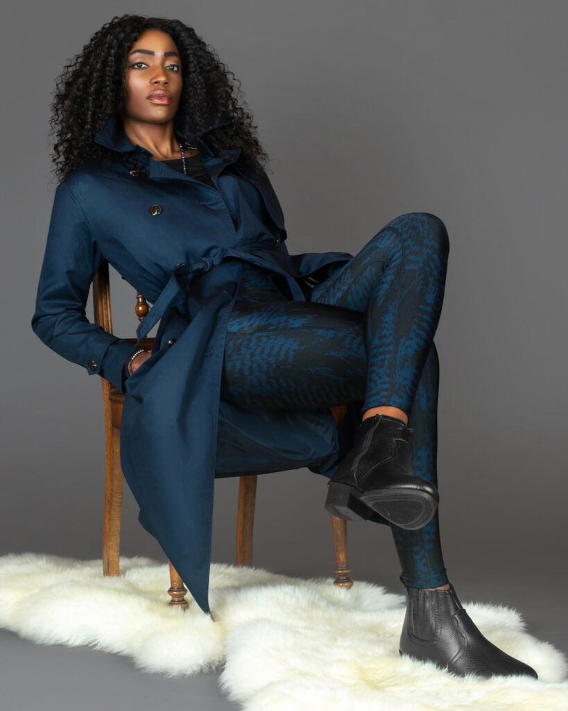 Portrait of a beautiful black woman with long curly black hair, beautiful makeup sitting by herself on fur in a studio with a grey background wearing a blue trench coat and leggings with black boots.