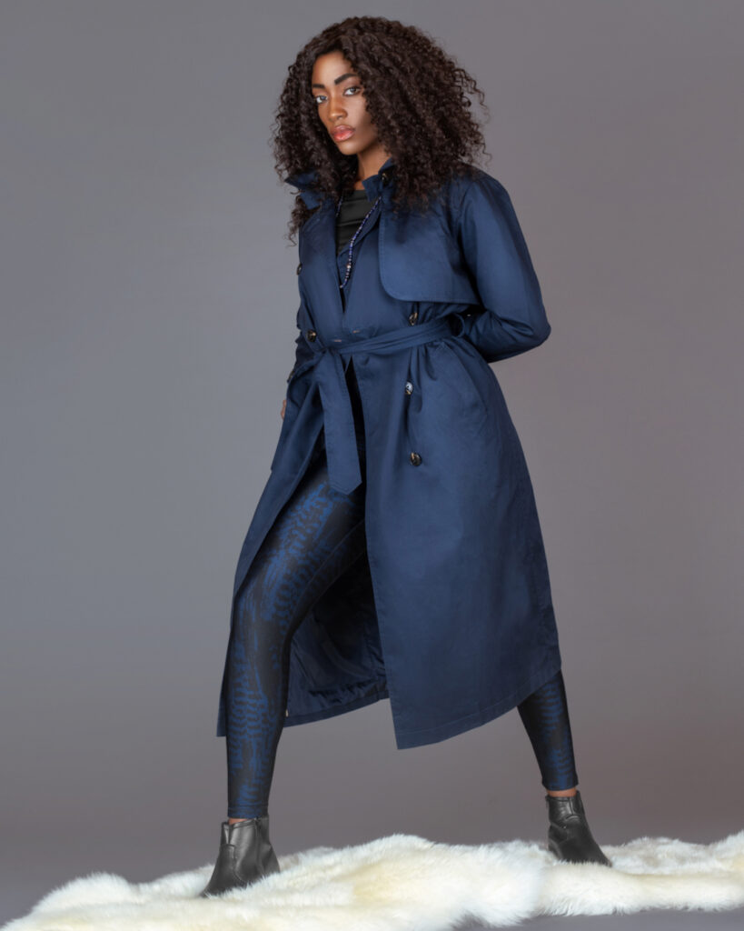 Spring Is In The Air. Black woman in blue trench coat, leggings and black boots sitting on fur in studio.