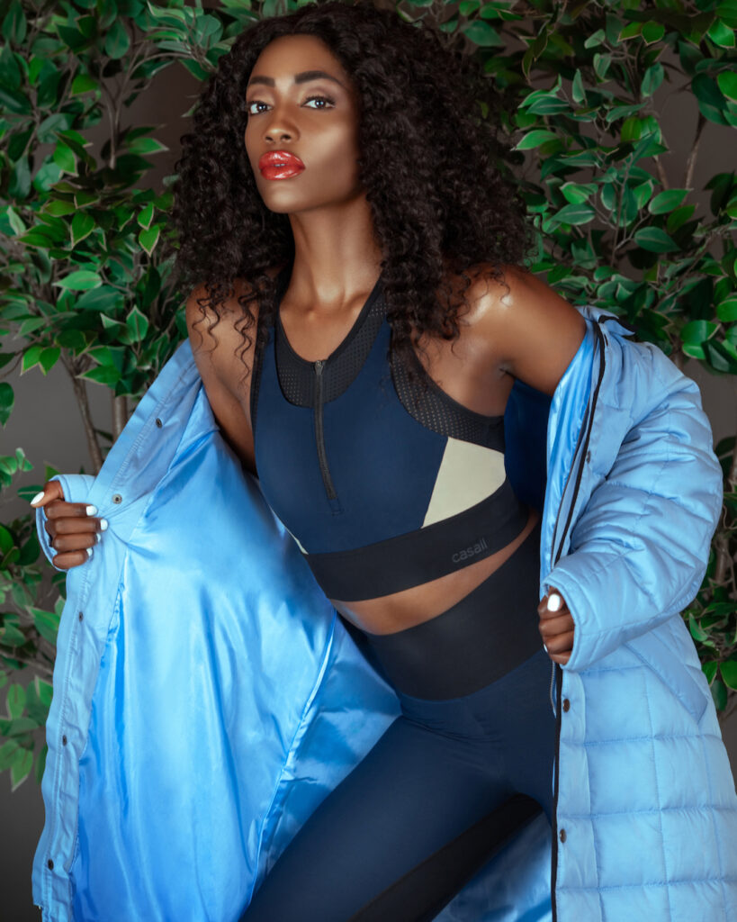 Spring Is In The Air. Black woman with curly hair in blue fitness outfit and light blue coat posing in front of green leaves background.